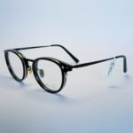 Black round eyeglasses with transparent lenses and metal bridge, displayed on a soft gradient background. Available at www.spexovision.com